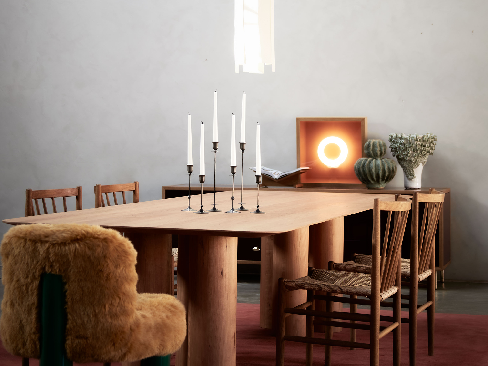 A wooden table with a candle holder on it, four wood chairs and a furry orange chair surrounding it. There is also a furniture piece in the background with a book, different vases and a circle lamp inside a wooden frame.