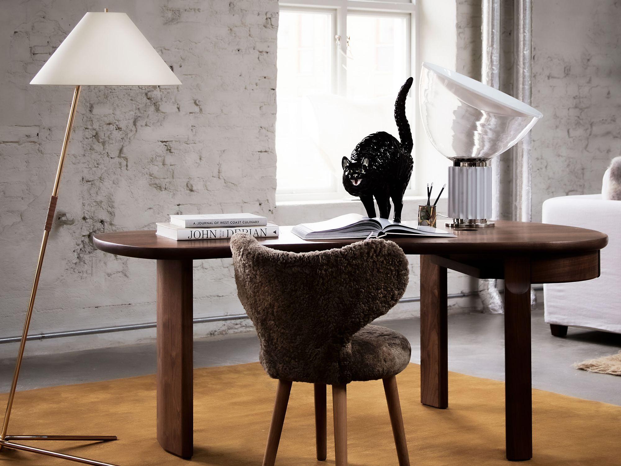 A dark wooden desk with some books, a lamp, a pencil holder, and a black cat sculpture on top of it. There is also a furry brown chair by the desk and floor lamp to the left of it.