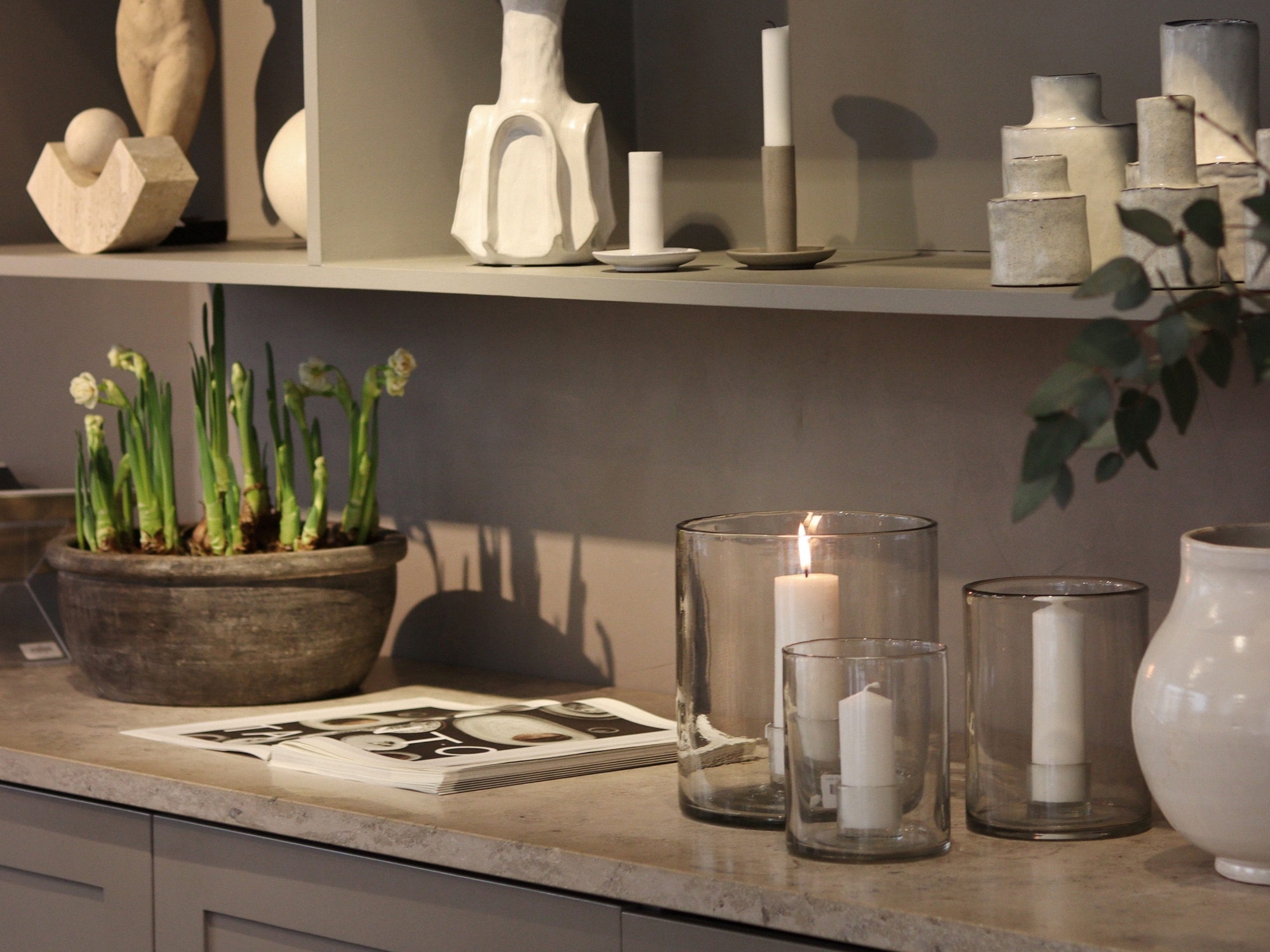 Different candle holders, such as ceramic and glass ones, a book opened on the counter and a vase with plants blooming.