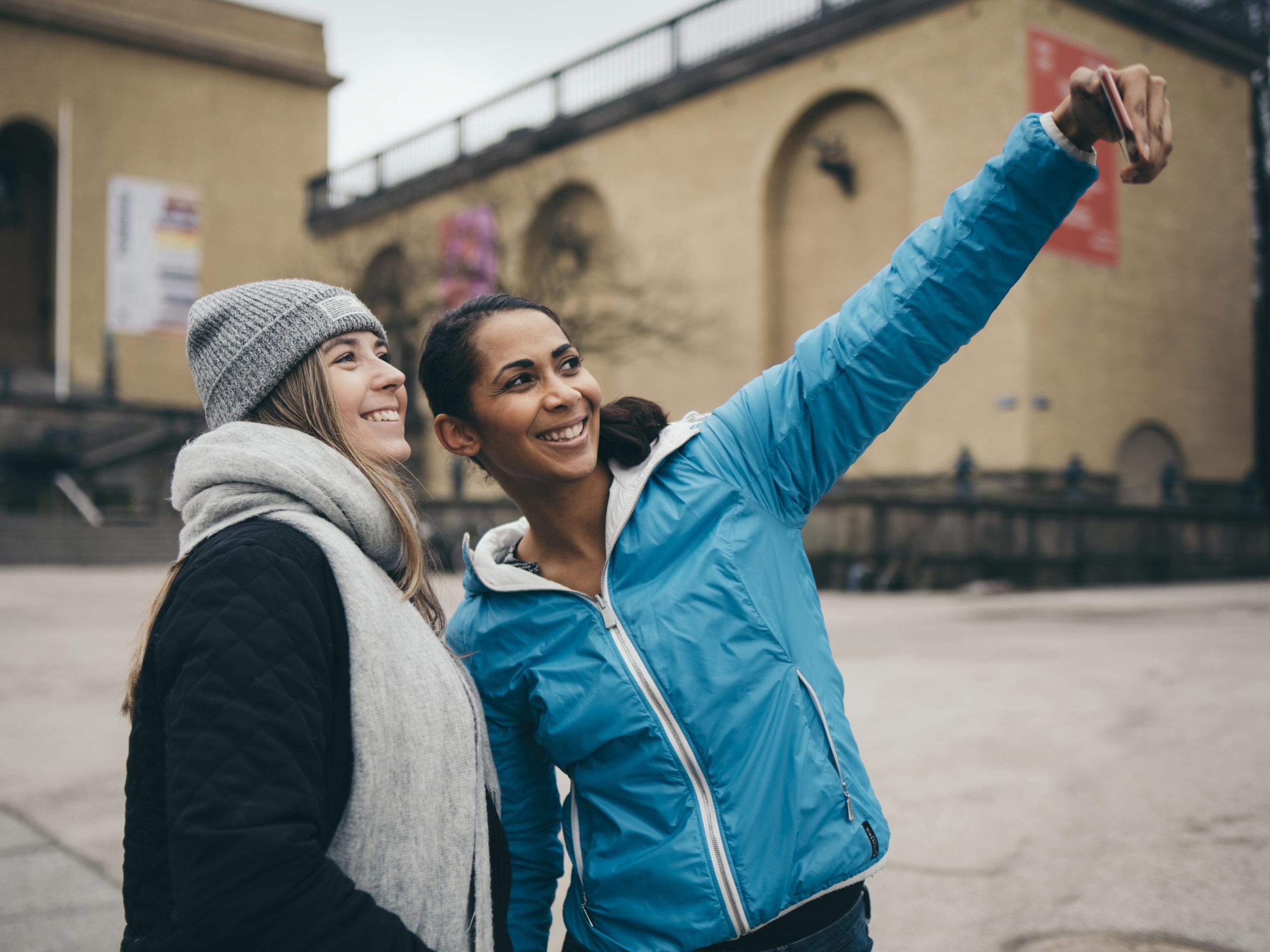 två tjejer i vinterkläder tar selfie på ett torg