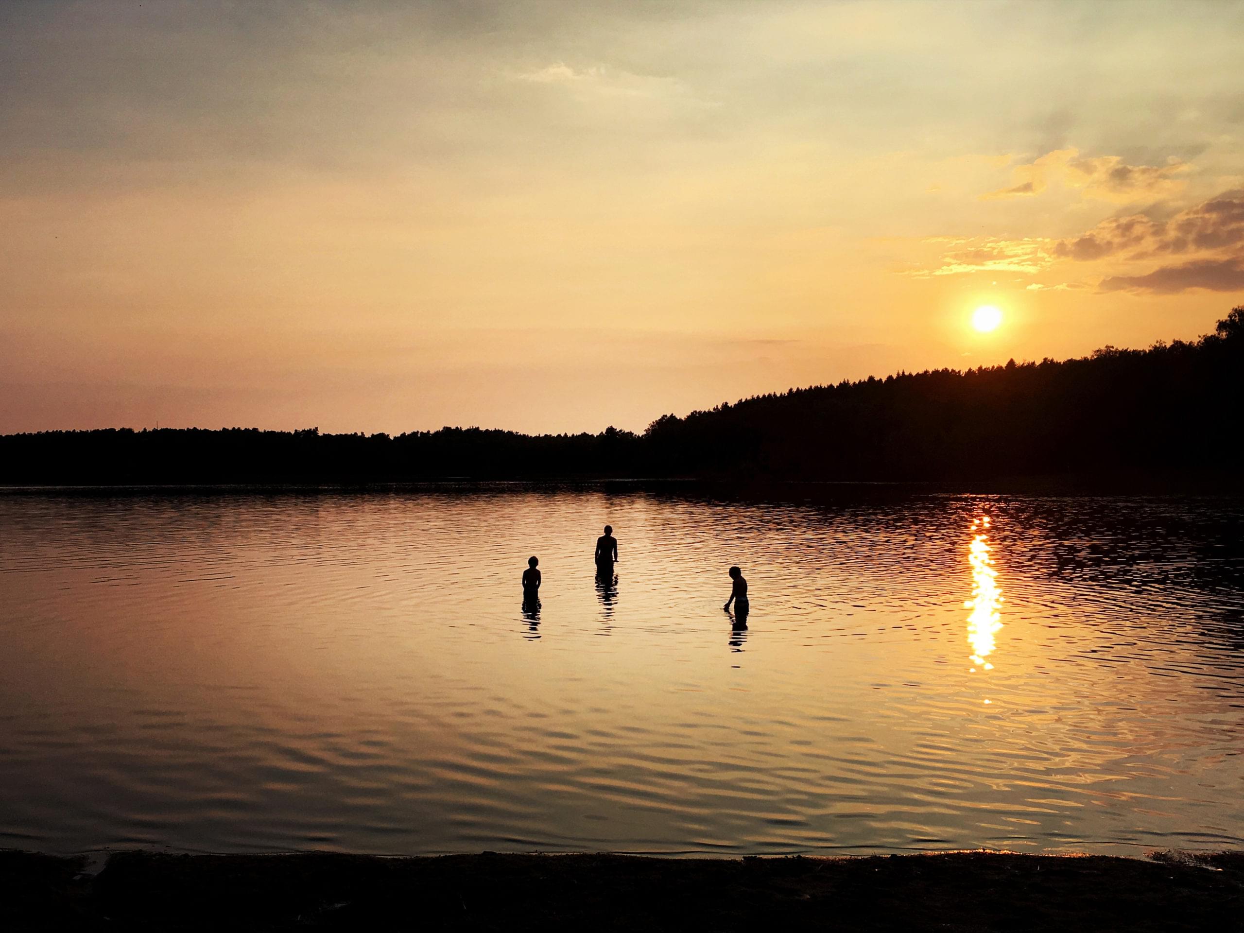 Tre personer bada i Härlanda Tjärn i kvällsljus.