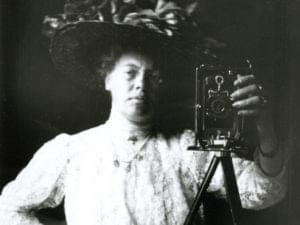 Ett svartvitt fotografi av en kvinna i stor hatt som fotograferar