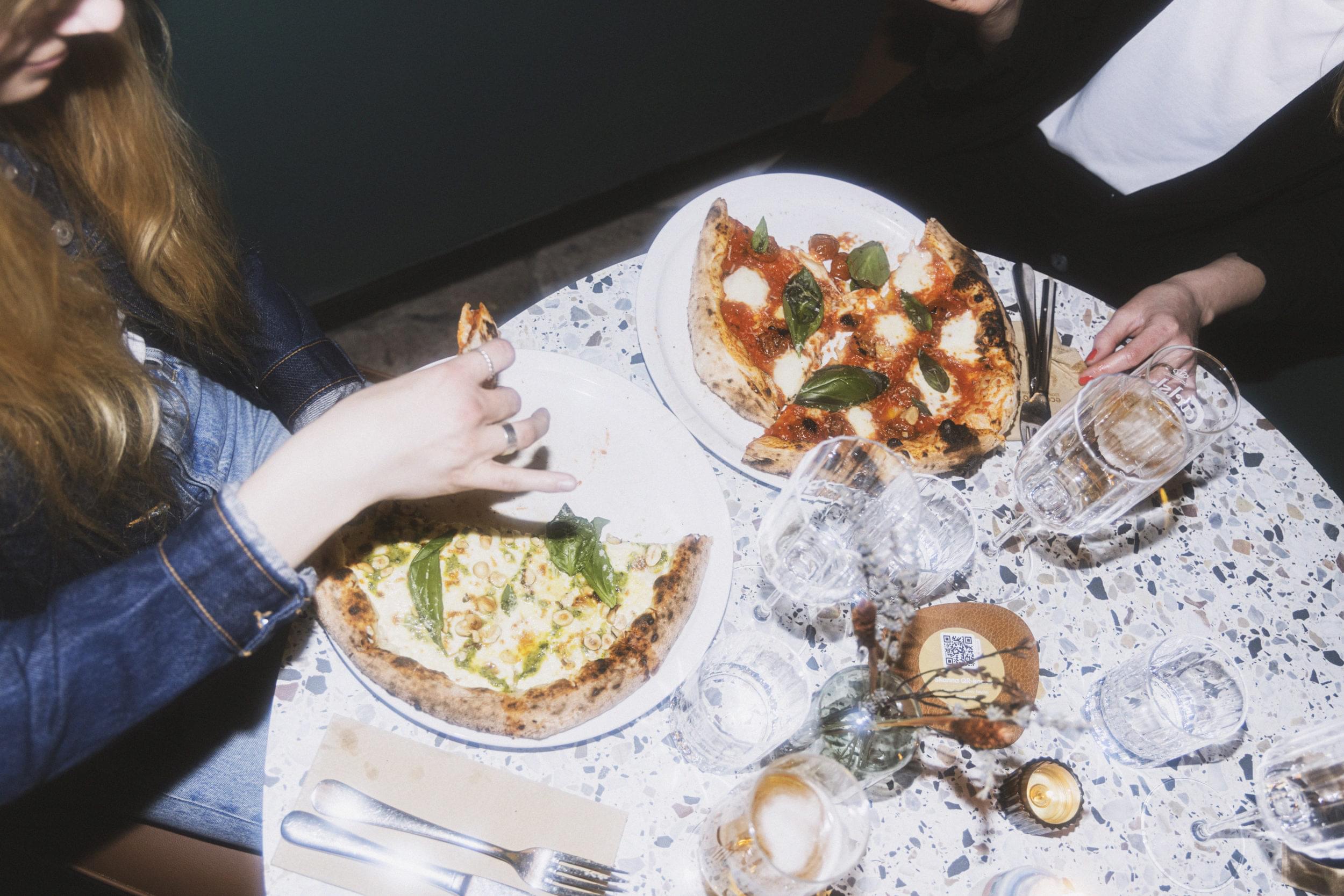 Runt, ljust bord med två pizzor på. Till vänster syns en hand som håller in en pizza slice.