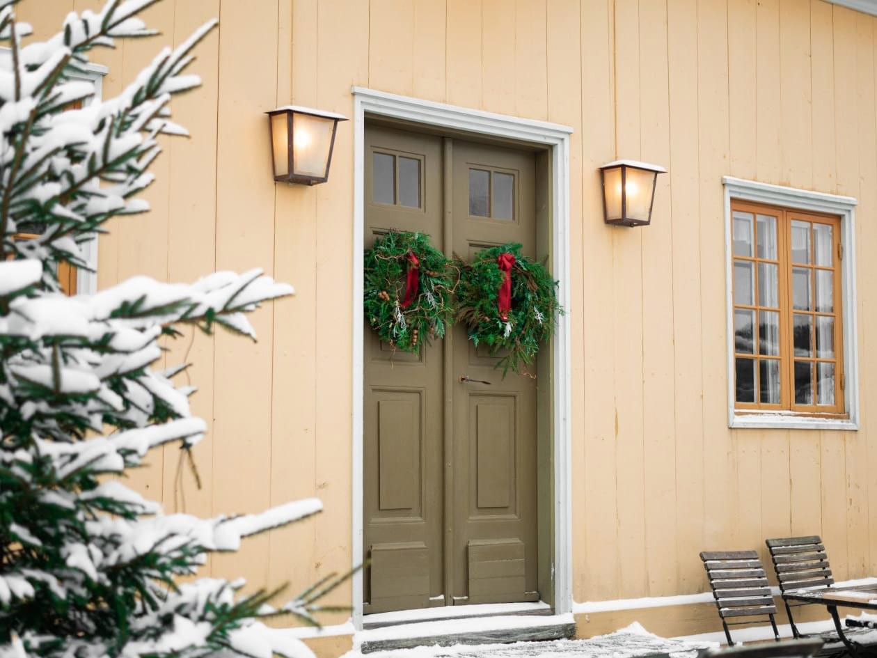En julgran klädd med snö står framför ett hus med en dubbeldörr. På dörrarna hänger två julkransar. 