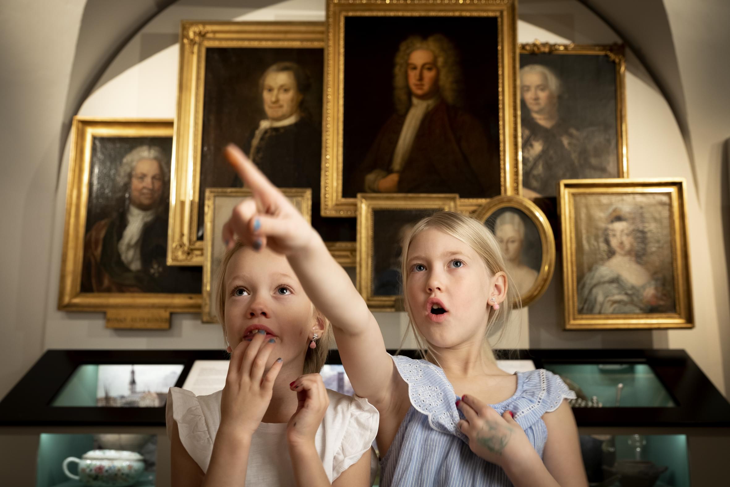 Två barn står framför en vägg av tavlor och porträtt av människor i 1700-talsperuker. De pekar på någonting de ser.