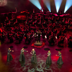 Sångare på scen i förgrunden, dirigent och orkestermusiker längre bak i bild.