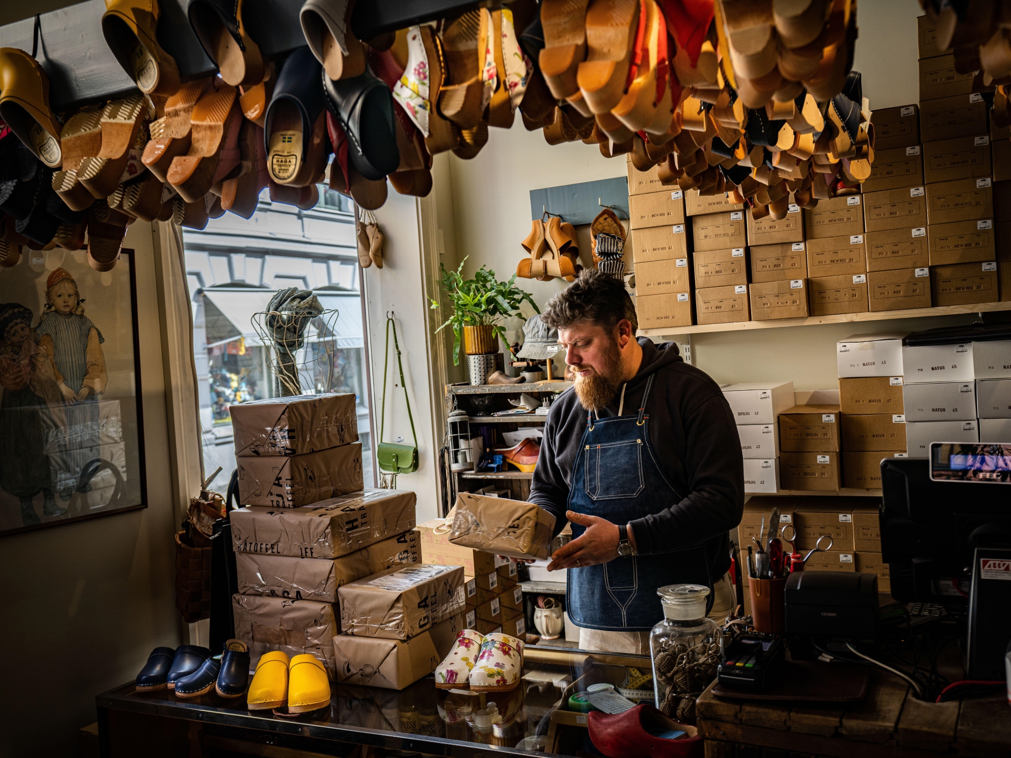 Man holding a package in a shop with different boxes and wooden clogs around him; mostly hanging from the ceiling.