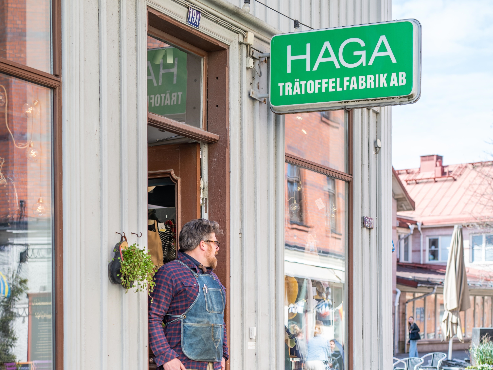 Butiksingång med en man som står vid dörren. Butiken har en grön skylt med "Haga Trätoffelfabrik AB" skrivet där.