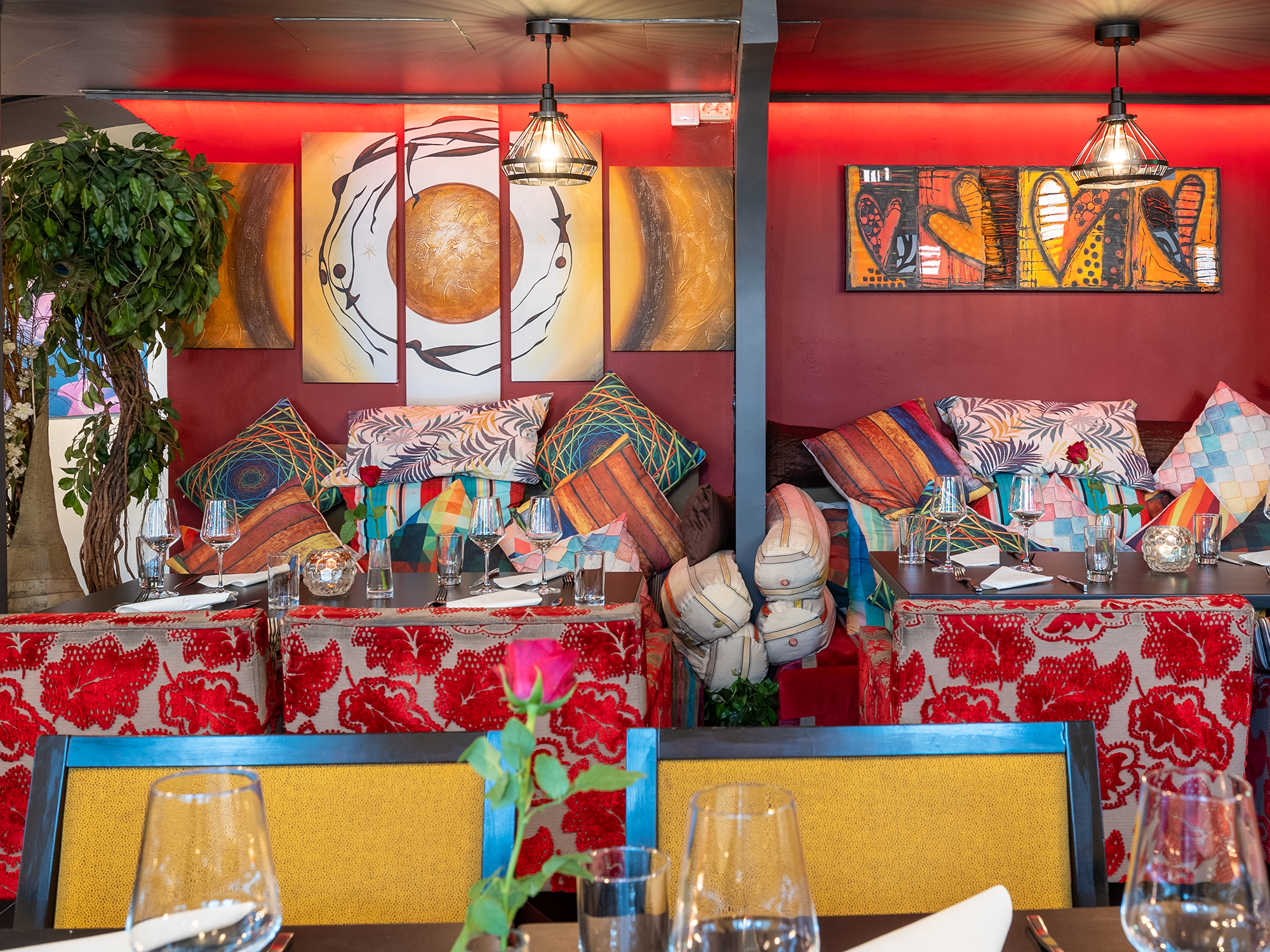 Restaurangsittmiljö med kuddar i olika färger och mönster, dukade bord och en trädplanta i rummets vänstra hörn