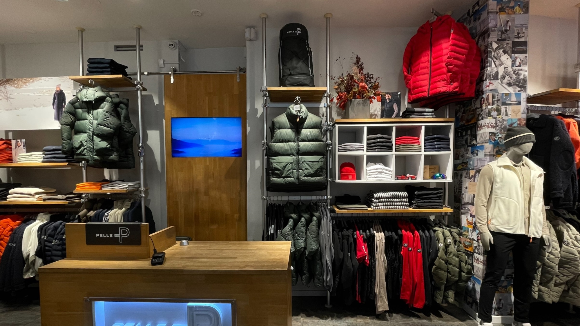Clothing store with different winter thick jackets, t-shirts, a backpack, and other clothing pieces. There is also the payment counter with a TV in the back and a mannequin on the right side.
