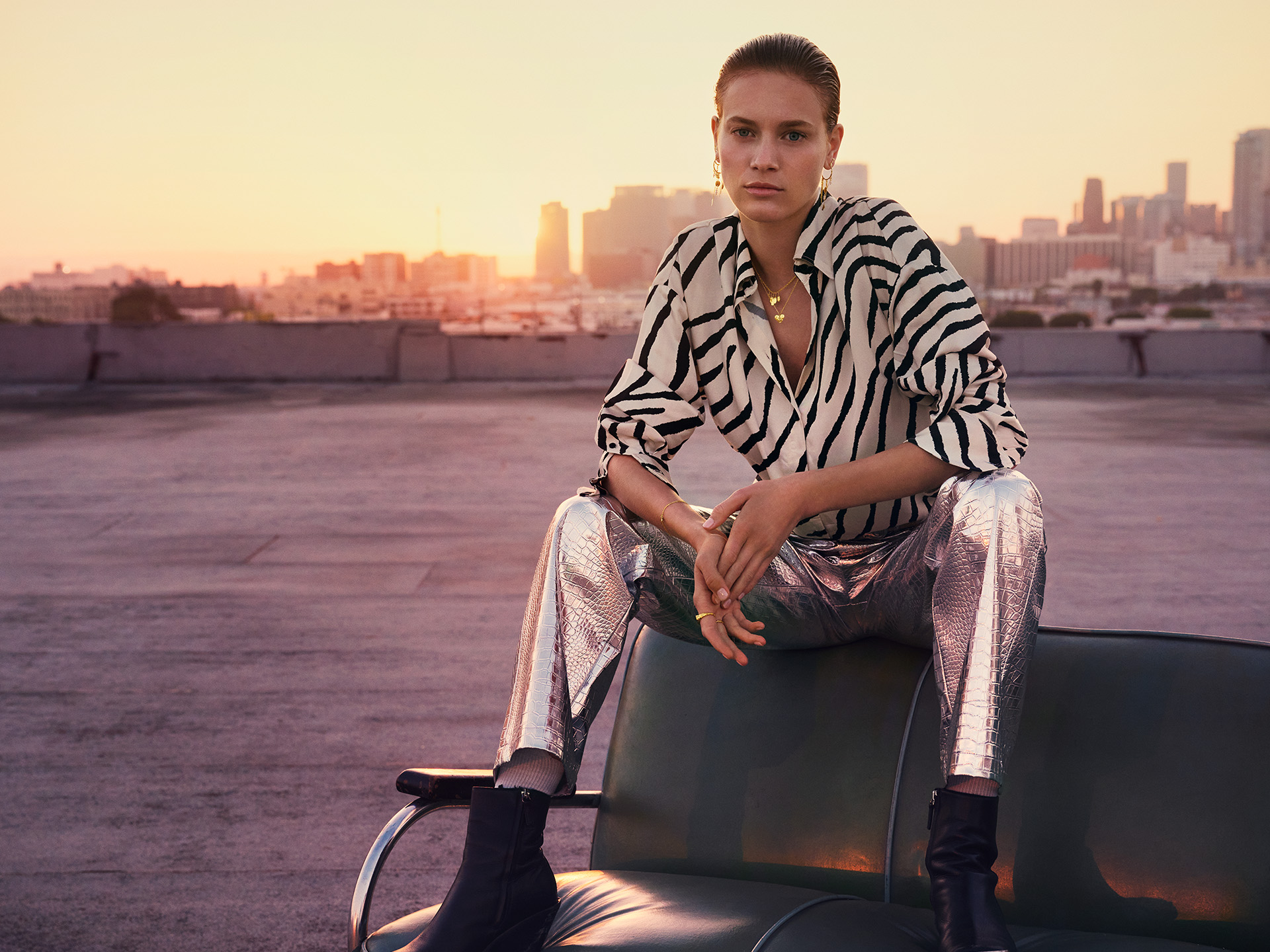 model sitting on top of a sofa using golden jewellery and using a zebra patterned blouse, crocodile textured silver pants and black boots.