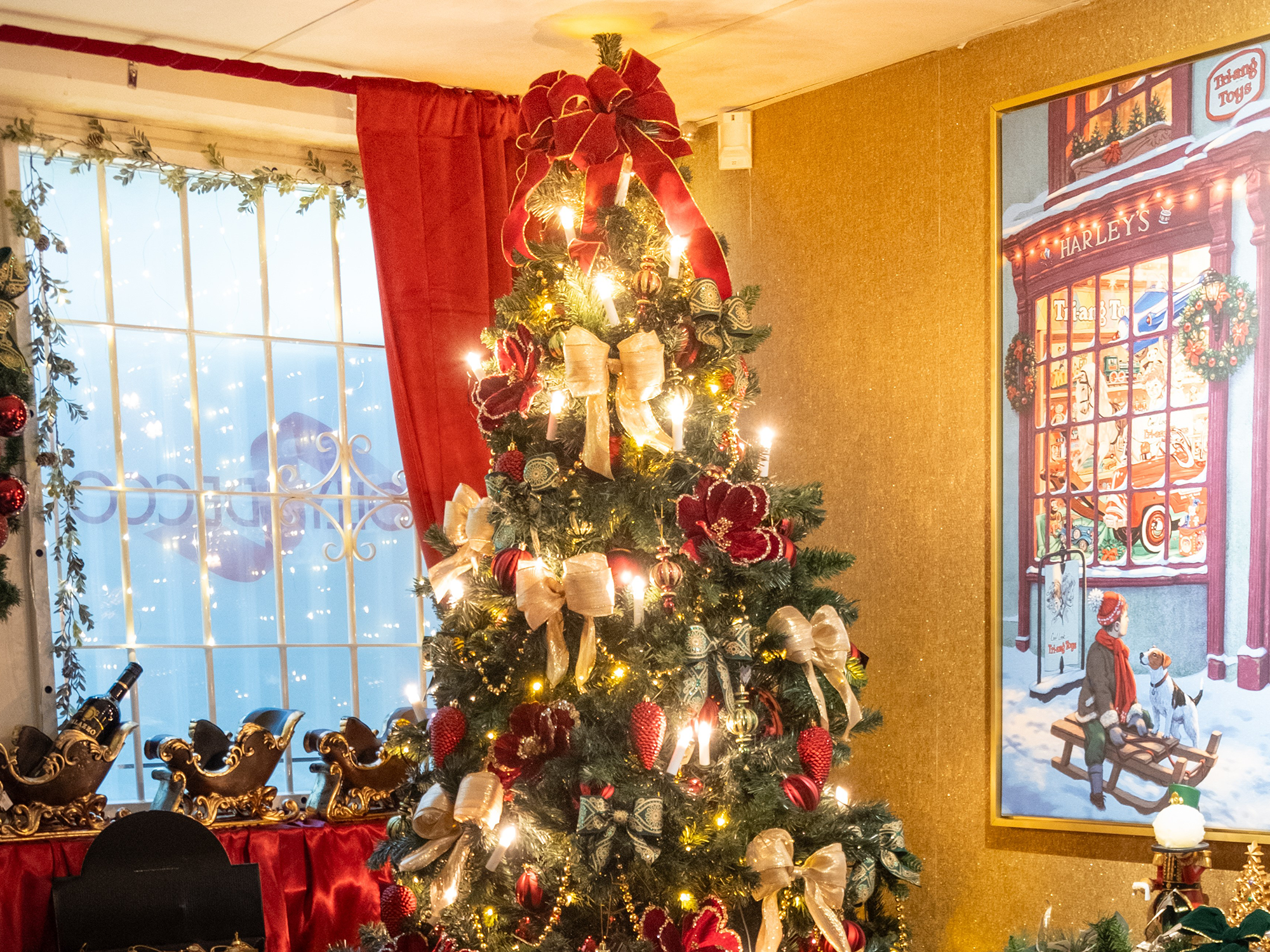 Julpyntat rum med en julgran i mitten, tre tomteslädar till vänster och en affisch med en pojke som sitter på en släde med en hund och tittar på ett julskyltfönster till höger.