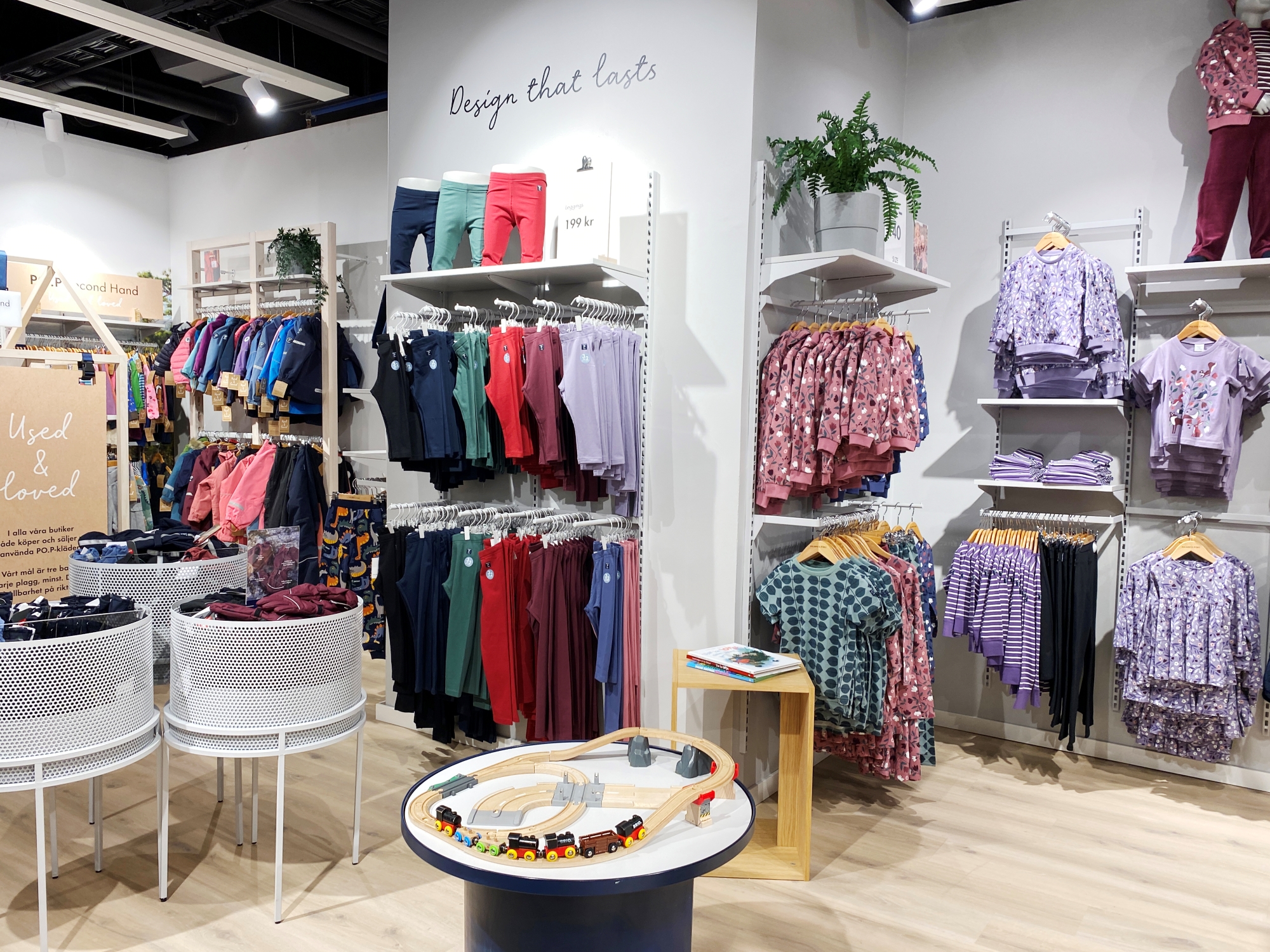 Colourful and patterned clothes for children hanging on a store. There is also a table with a wooden track and cars on it in the middle of the store and a quote on the wall that says, “design that lasts”.